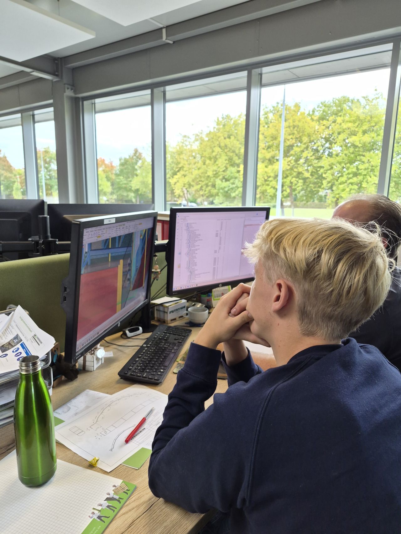 Een jonge student bekijkt aan een bureau met twee schermen een 3D ontwerp van een machine en een onderdelenlijst, met een technische tekening op het bureau.